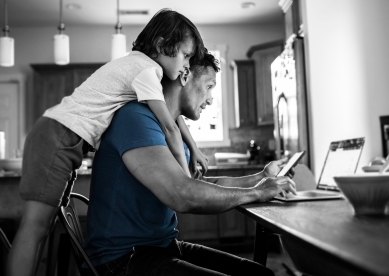 Son hanging on the back of his dad, while the dad uses a laptop in the kitchen of their house